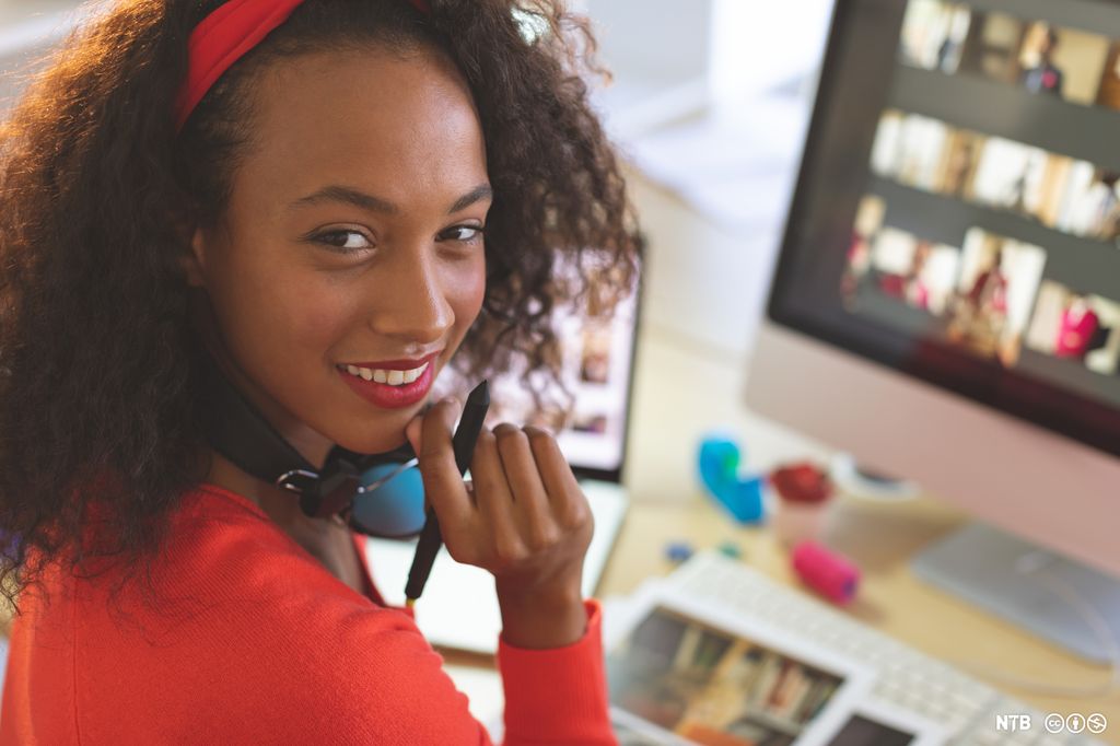 Ung kvinne med krøllete hår og rød genser ser mot kameraet. I bakgrunnen en datamaskin med flere bilder på. Foto.