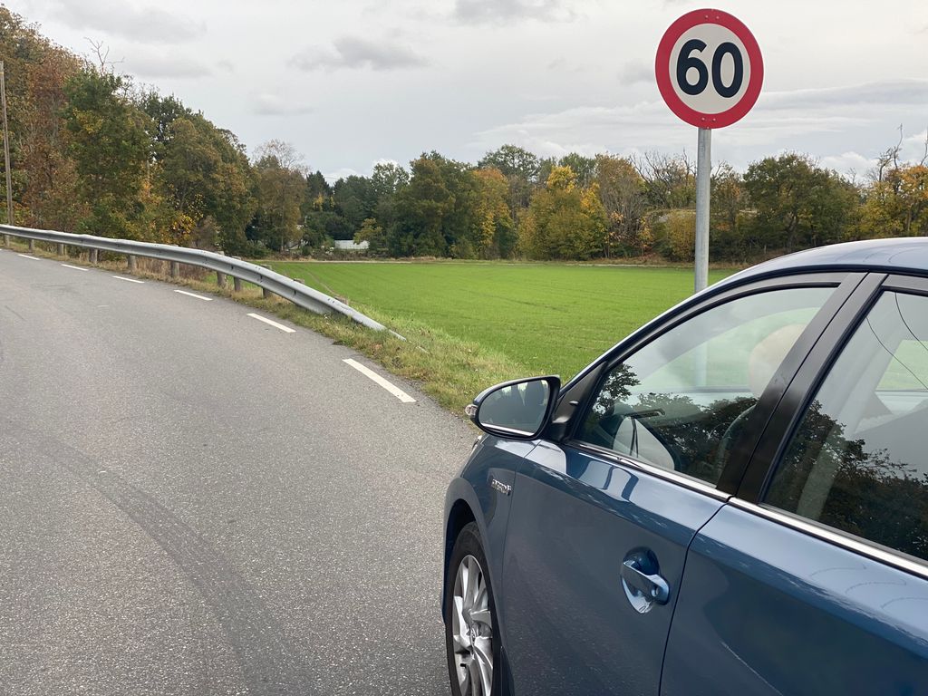 Blå personbil som står på høyre side i veibanen. Bilen står like før et skilt som viser 60-sone, og veien framover svinger mot venstre. Foto. 