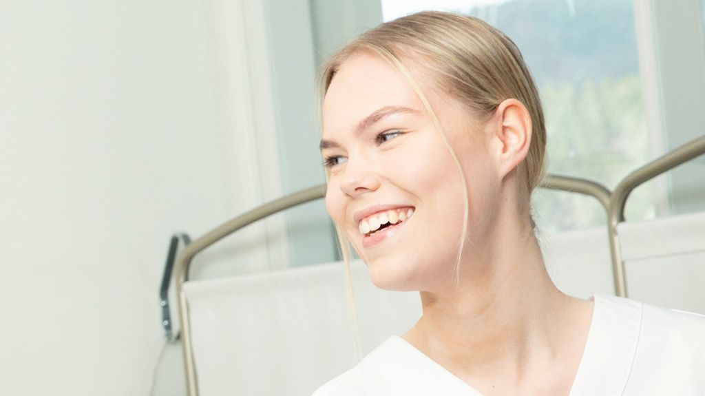 Helsefagarbeider i hvit uniform smiler bredt. Foto.