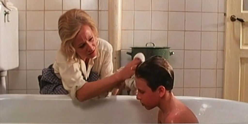 Ein gut sit i badekaret. Ei kvinne sit bøygd på golvet ved badekaret og vaskar håret hans. Foto. 