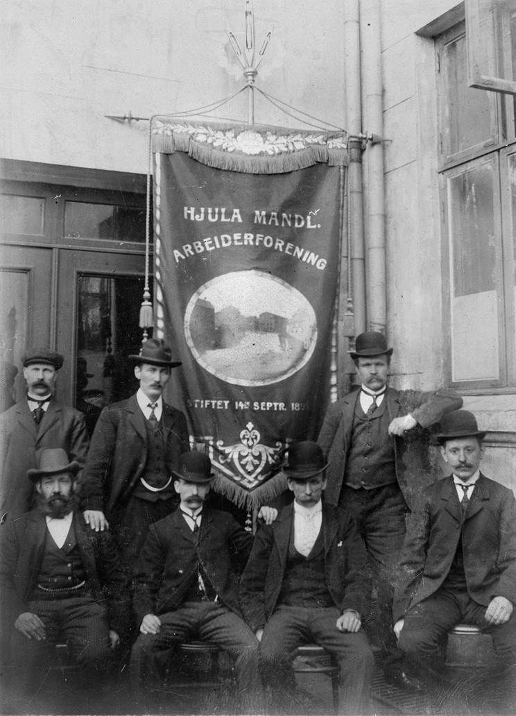 Sju menn i dress og med hatt. Fire av dem sitter på stoler, de tre andre står bak med en  fane som det står "Hjula mandl. arbeiderforening" på. Svart-hvitt foto.