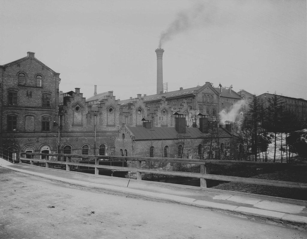 Fabrikkbygninger med ei høy pipe det kommer røyk fra. Bildet ser ut til å være tatt ved en bro, og vi ser ei elv i bakgrunnen. Bildet er i svart-hvitt. Foto. 