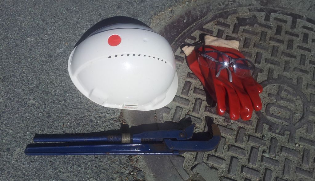 Hjelm, hansker, vernebriller og rørtang på kumlokk. Foto.