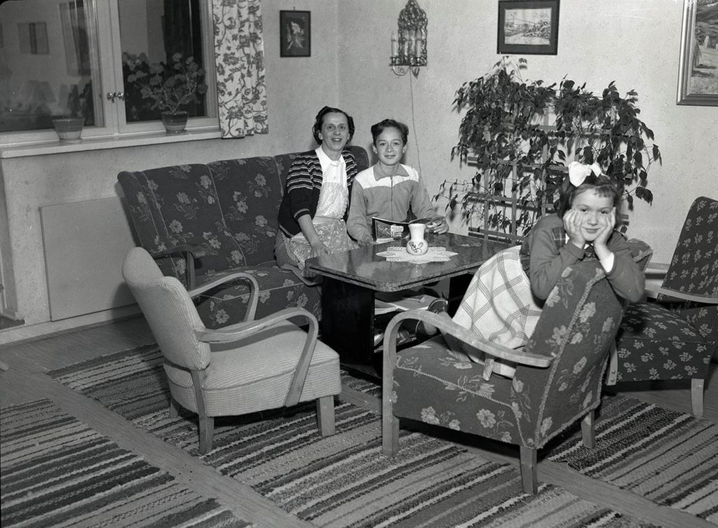 Ei husmor og to barn i ei sitjegruppe i ei stove på 1950-talet. Foto.