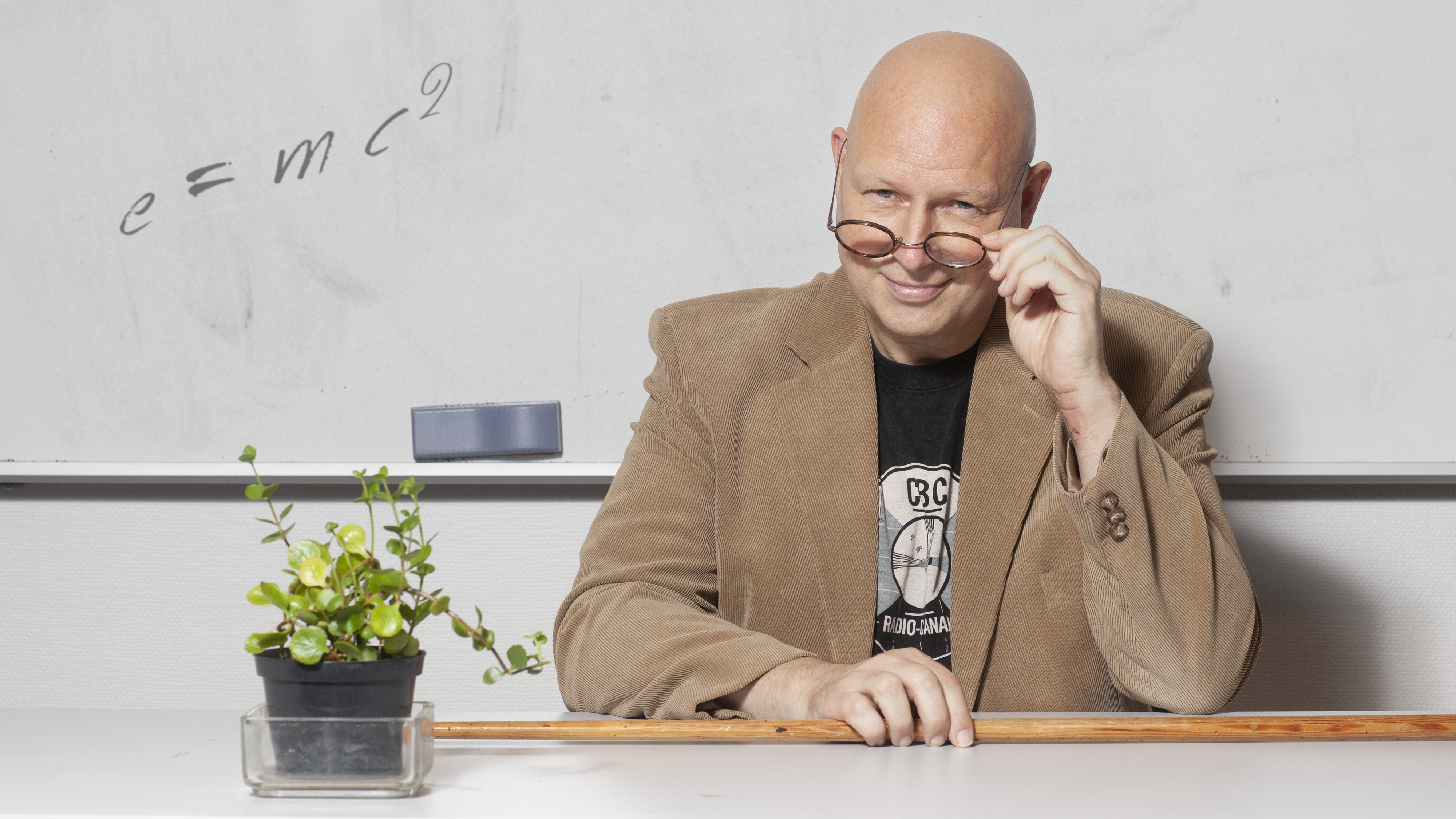 En smilende mann sitter og holder brillene ned på nesa. Han sitter ved et kateter foran ei hvit tavle. Foto.