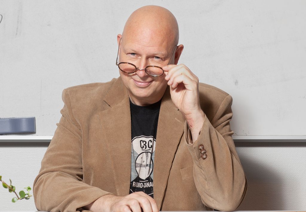 En smilende mann sitter og holder brillene ned på nesa. Han sitter ved et kateter foran ei hvit tavle. Foto.