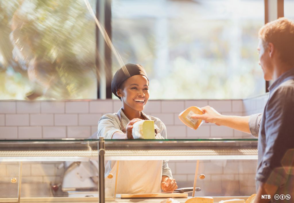 En kvinnelig ferskvarehandler smiler til kunden mens hun legger ost på disken. Foto.