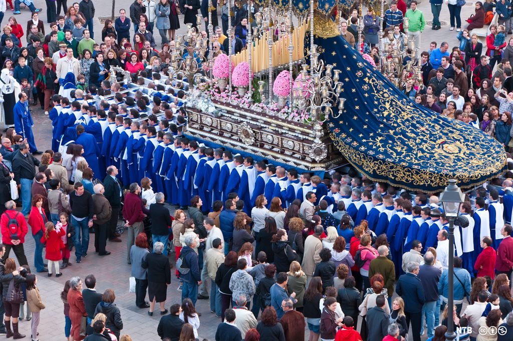 Ei flåte pyntet med lys, blomster og en statue av jomfru Maria bæres gjennom gatene. Det er mange tilskuere rundt. Foto.