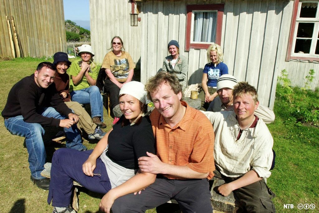 Ei gruppe kvinner og menn i fritidsklede framfor eit lite gardsbruk. Foto.