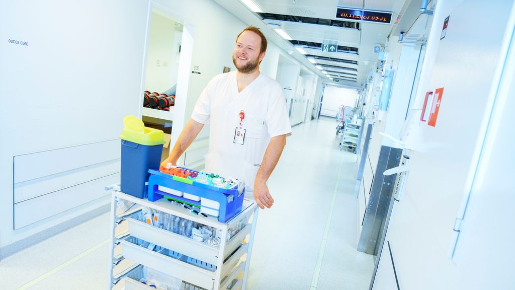 Person i uniform for helsepersonell skyver tralle med medisinsk utstyr i en korridor. Foto.