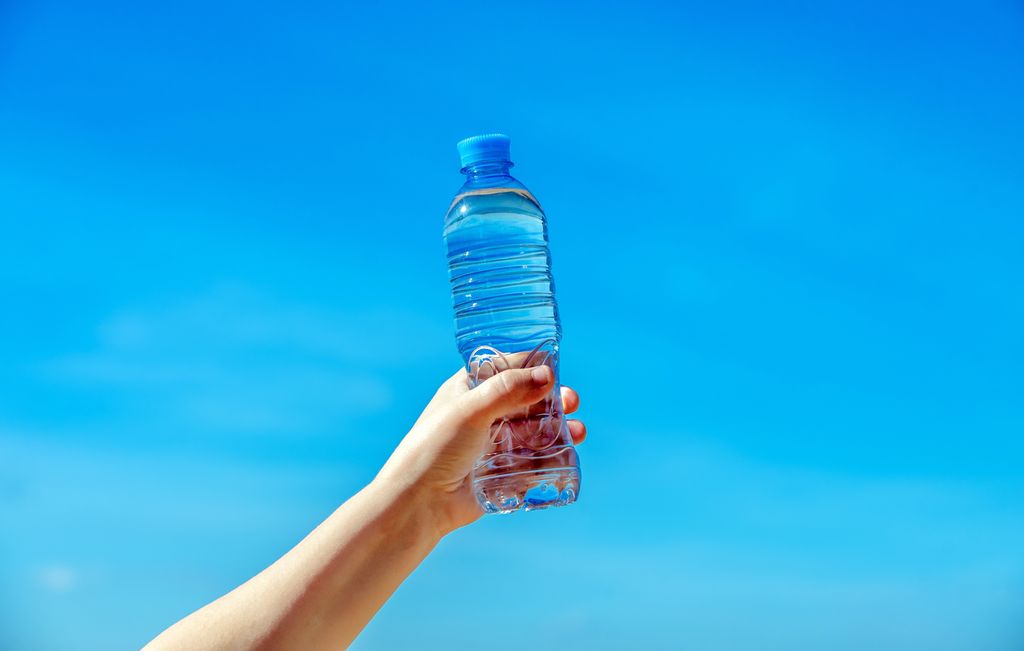 Ei hånd holder ei flaske vann mot en blå himmel. Foto.