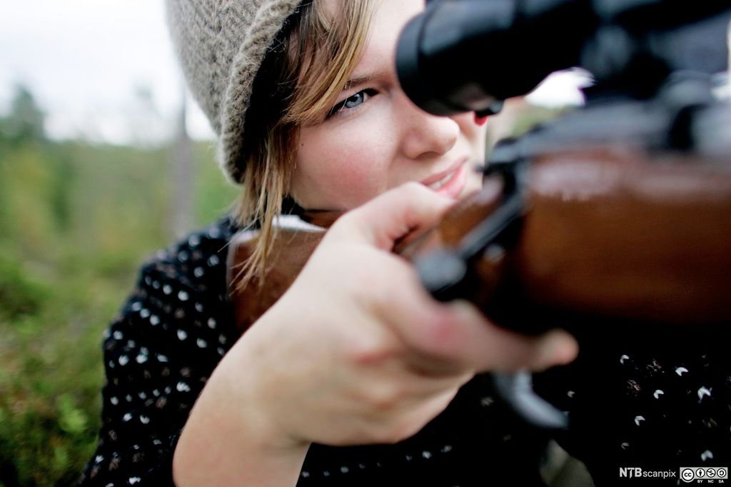 Ung person i naturen som sikter gjennom et kikkertsikte på et gevær, med fokusert uttrykk. Personen har på seg en strikkelue og mørk genser, og bakgrunnen viser et grønt skogslandskap. Foto.