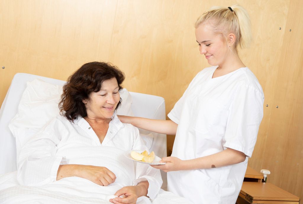 Pasient i seng får servert frukt av helsefagarbeider. Foto.