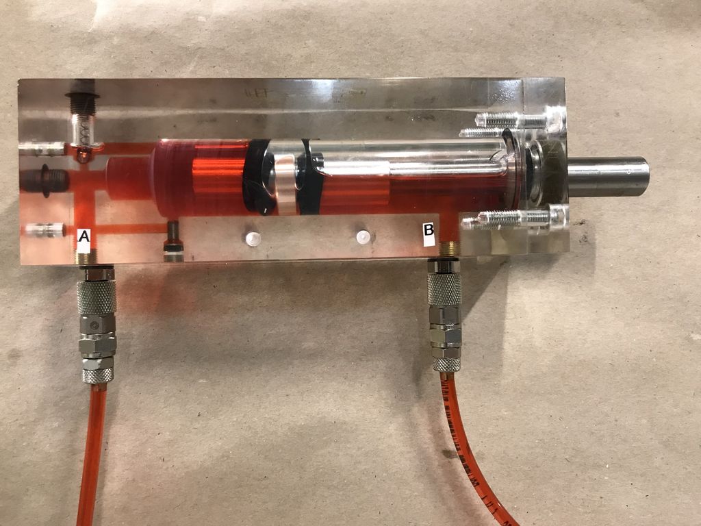 En glassylinder som er festa til en metallbit, som igjen er festa til en vegg. Sylinderen er delvis fylt med væske. Inne i sylinderen er det også et såkalt stempel, det vil si en metallpinne med ei rund plate i den ene enden. Det er kopla til to vannledninger, ei i hver ende av sylinderen. Foto.