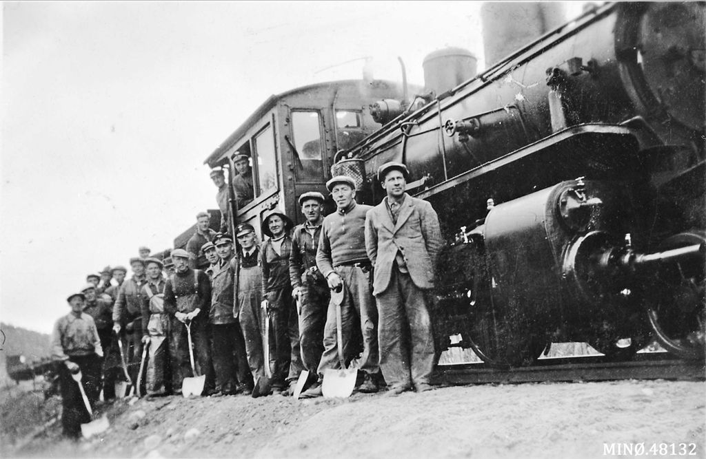 Menn med spader og hakker i hendene foran et stort damplokomotiv. Svart-hvitt-foto.