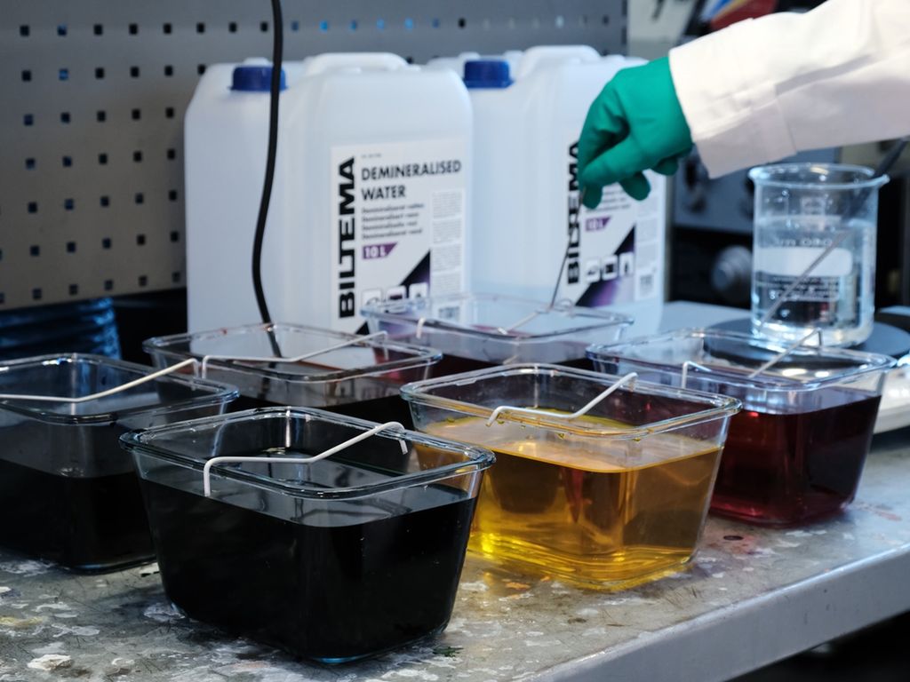 Firkanta glassbeholdere på en arbeidsbenk. De inneholder væske av ulik farge, blant annet gult og rødt. Foto.