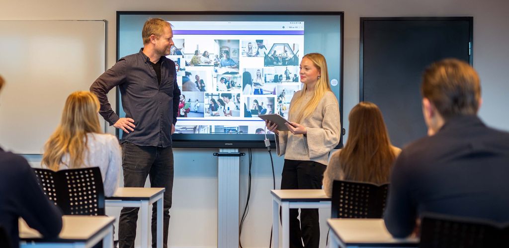 Klasserom hvor noen elever sitter og en elev står sammen med lærer oppe ved tavla.