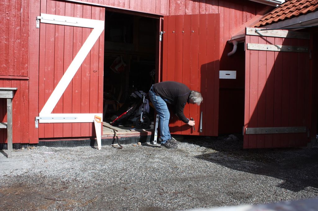 En mann står bøyd mot ei åpen garasjedør og jobber med verktøy. Ei sag og et vaterpass står bak ham. Foto.