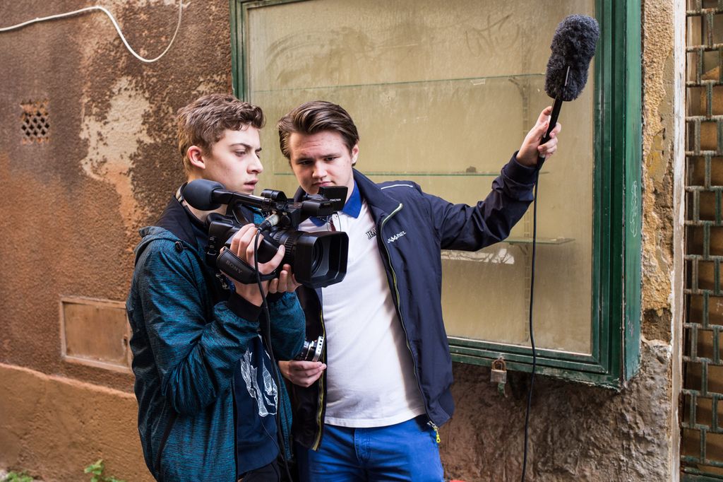 To gutar står framfor ein murvegg. Den eine filmar med eit videokamera, den andre held ein mikrofon opp i vêret. Foto.