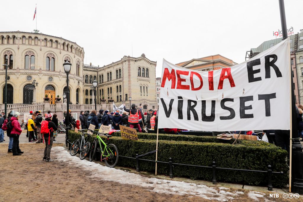 En gruppe mennesker med plakater samlet foran Stortinget. I forgrunnen et stort banner med teksten "media er viruset". Foto.