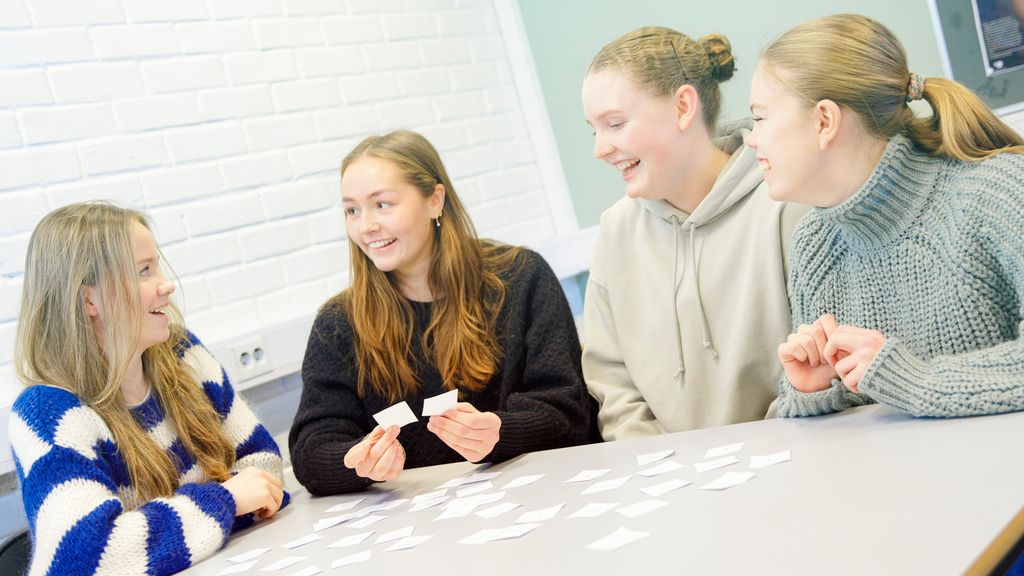 Fire jenter sitter ved et bord med mange små papirlapper på. Foto.