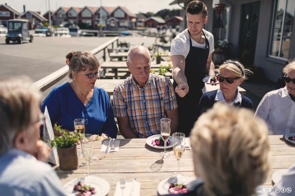 En ung servitør serverer mat ved et bord med eldre gjester på en uteservering en sommerdag. Foto.