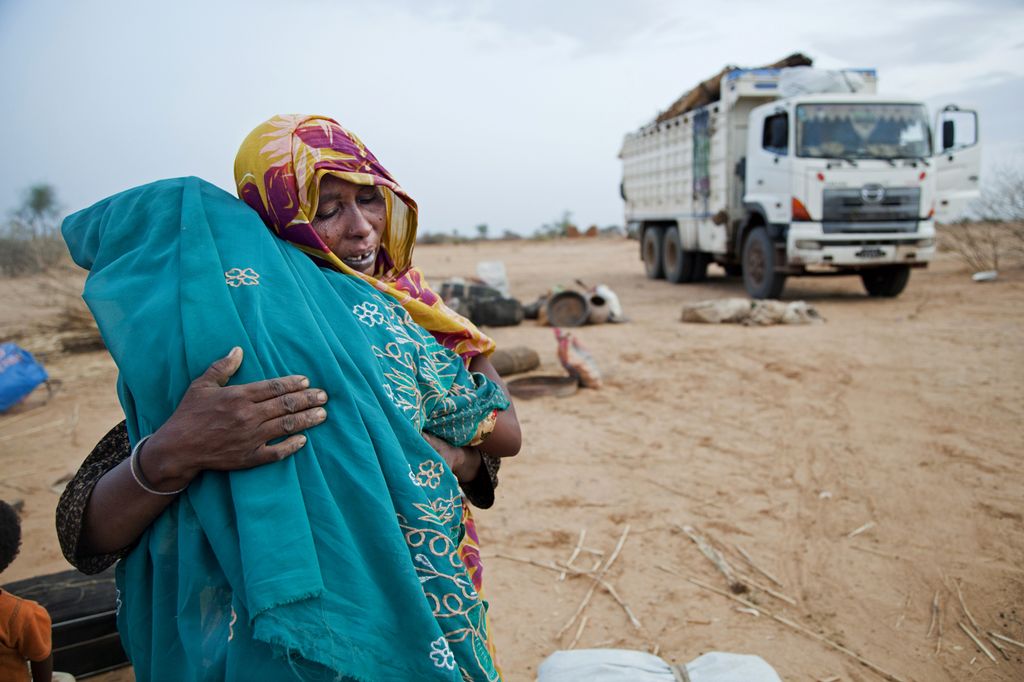 A woman wearing a yellow and purple headscarf is hugging a girl wearing a light blue headscarf. In the background there is a truck. They are in a sandy landscape. Photo.