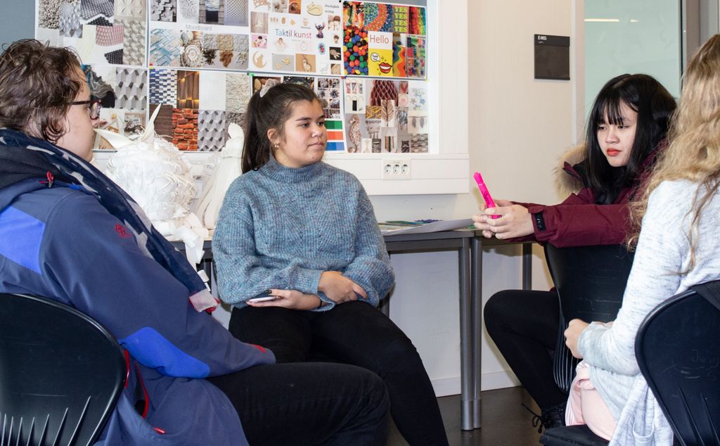 Fire elever sitter i ring i et klasserom, og en av dem holder en rosa penn. Elevarbeider henger på veggen bak. Foto.