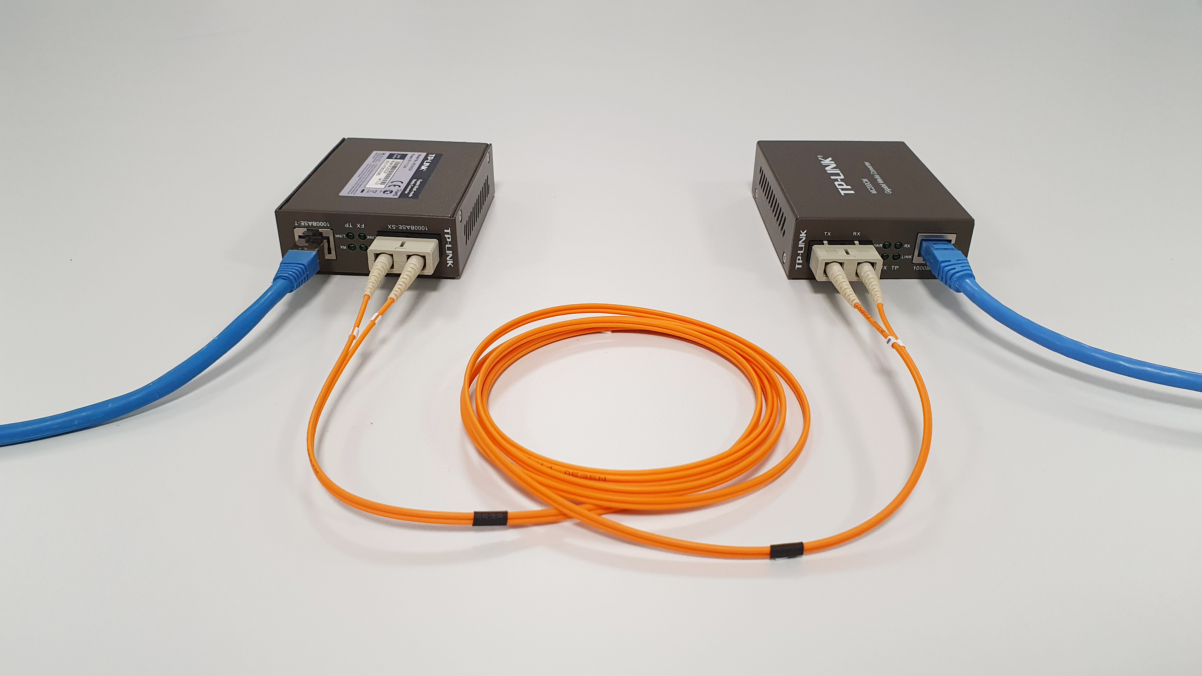 To små metallbokser koblet sammen med fiberkabel. Boksene har også hver sin nettverkskabel tilkoblet som går i hver sin retning ut av bildet. Foto.