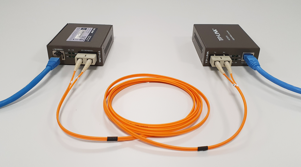 To små metallbokser koblet sammen med fiberkabel. Boksene har også hver sin nettverkskabel tilkoblet som går i hver sin retning ut av bildet. Foto.