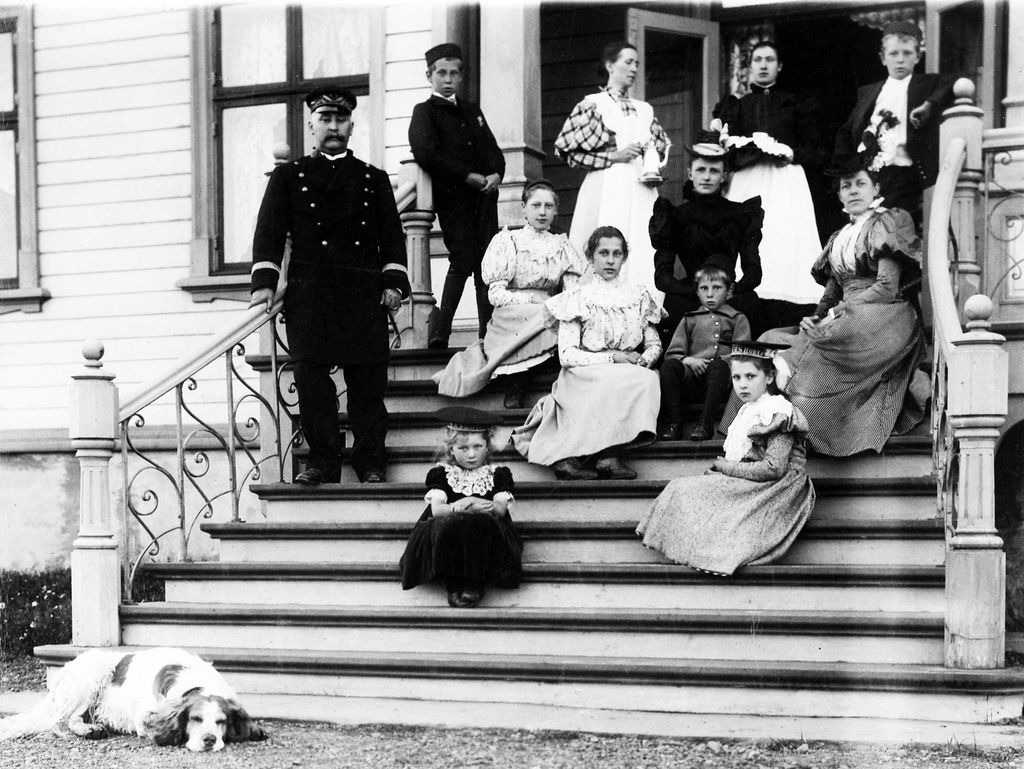 En familie står og sitter på trappen til et hus. Familien er kledd i finere klær. Noen på bildet er sannsynligvis hushjelper. Det ligger en hund nedenfor trappen. Huset ser stort ut, og trappen har flere trinn med forseggjort rekkverk. Bildet er fra Vardø i 1898 og viser amtmann Graff med familie og hushjelper. Svart-hvitt-foto.