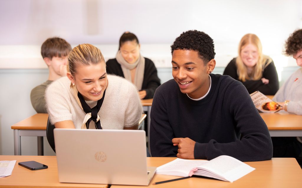 To elever sitter ved siden av hverandre i et klasserom og smiler mens de ser på en pc-skjerm. Foto.
