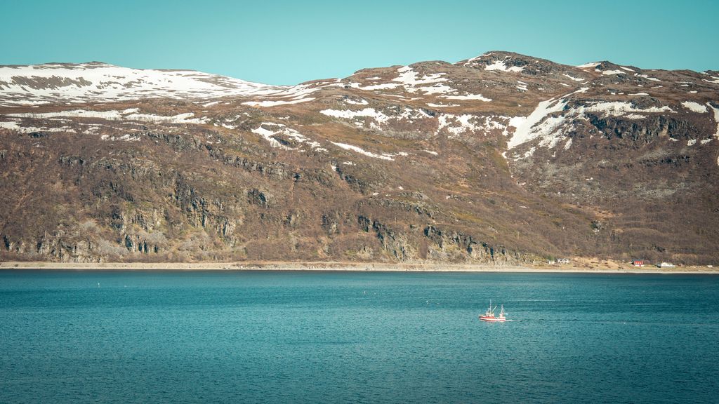 En fiskebåt på en fjord. Snødekte fjell i bakgrunnen. Foto.