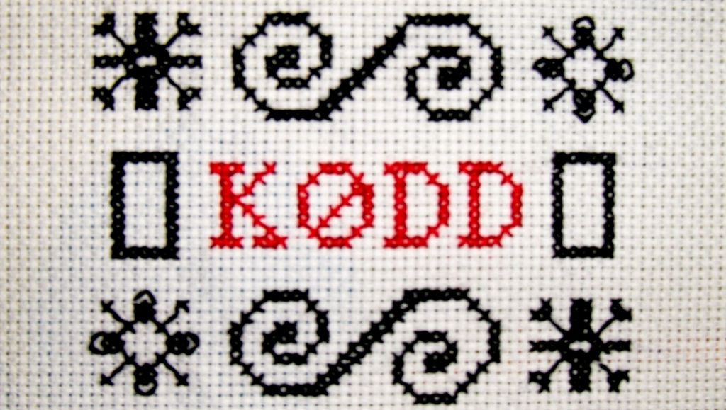 Et broderi der ordet KØDD i rødt og med store bokstaver er innrammet av ulike broderisting i svart. Foto.