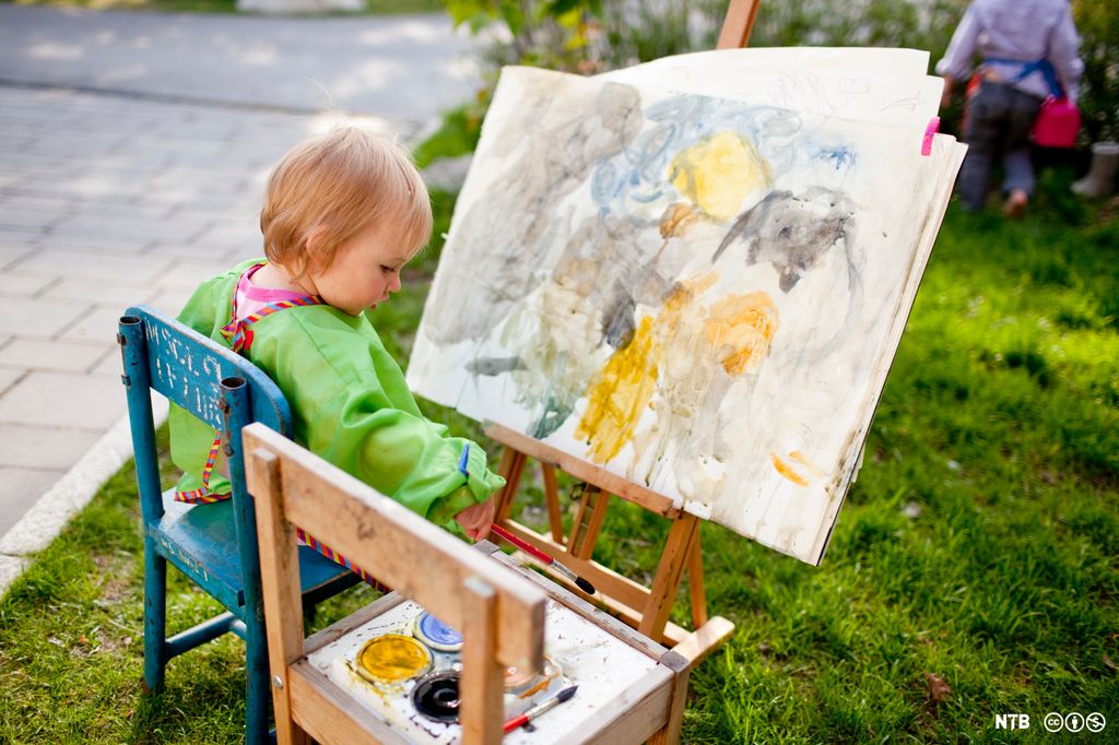 Lita jente sitter på en stol og maler på et lerret på et staffeli utendørs. Foto.