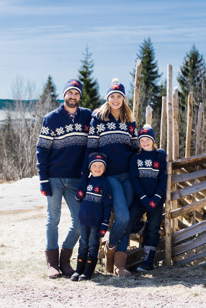 To vaksne og to barn som har på seg mørkeblå strikkegenserar og luer og vottar i same design. Alle smiler. Dei står ved ein skigard, og det er tre og snø i bakgrunnen. Foto.
