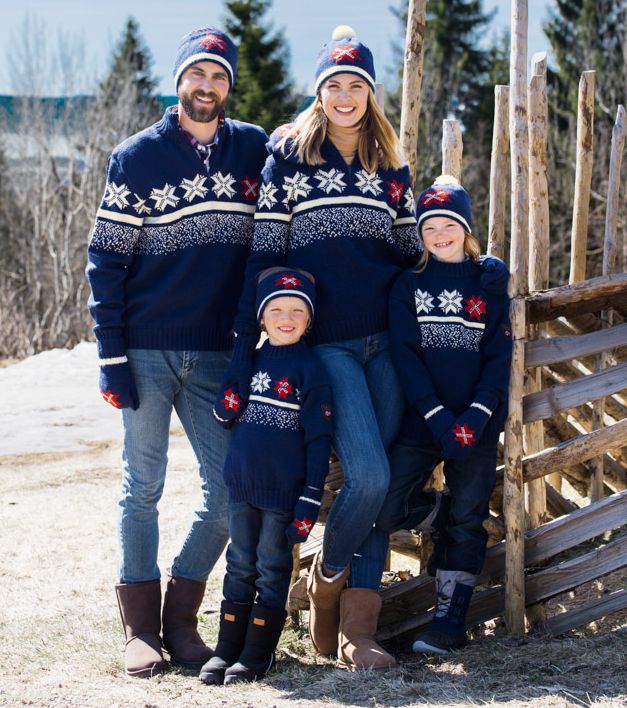 To vaksne og to barn som har på seg mørkeblå strikkegenserar og luer og vottar i same design. Alle smiler. Dei står ved ein skigard, og det er tre og snø i bakgrunnen. Foto.