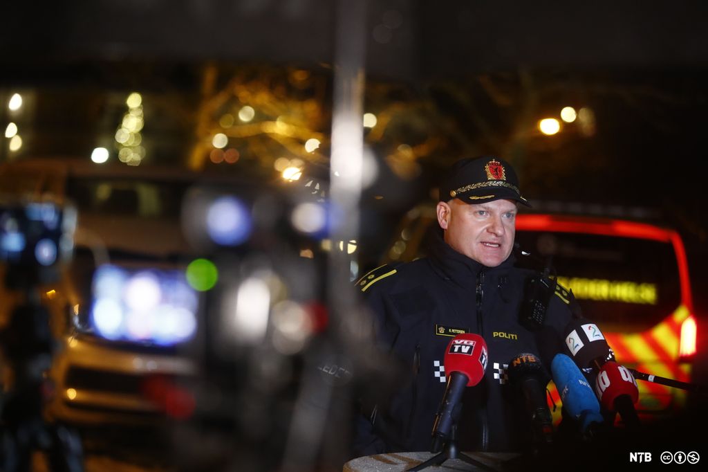 En mann i politiuniform står foran et bord med mikrofoner fra ulike norske medier. I bakgrunnen, som er ute av fokus, ser vi lys og utrykningskjøretøy. Foto.