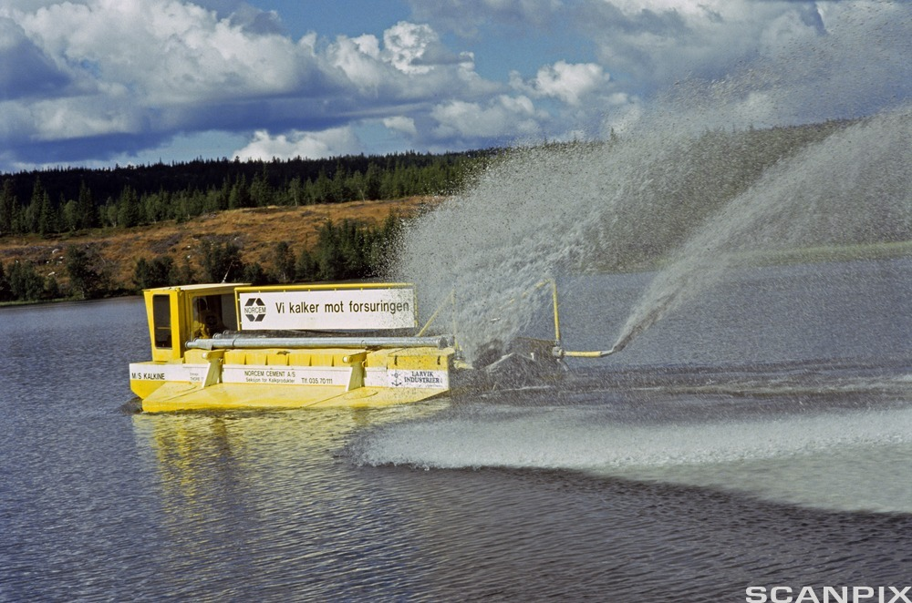 Gul båt sprer kalk på vann. Båten har et skilt der det står: "Vi kalker mot forsuringen". Foto.