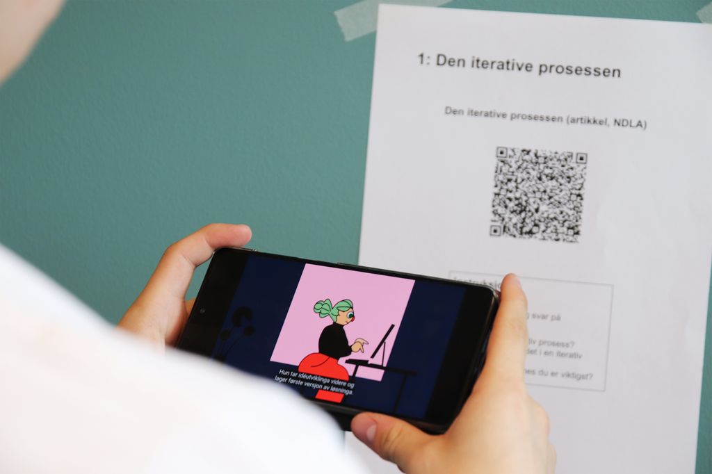 En person ser en video på mobilen. På veggen i bakgrunnen henger det et ark med QR-kode. Foto.