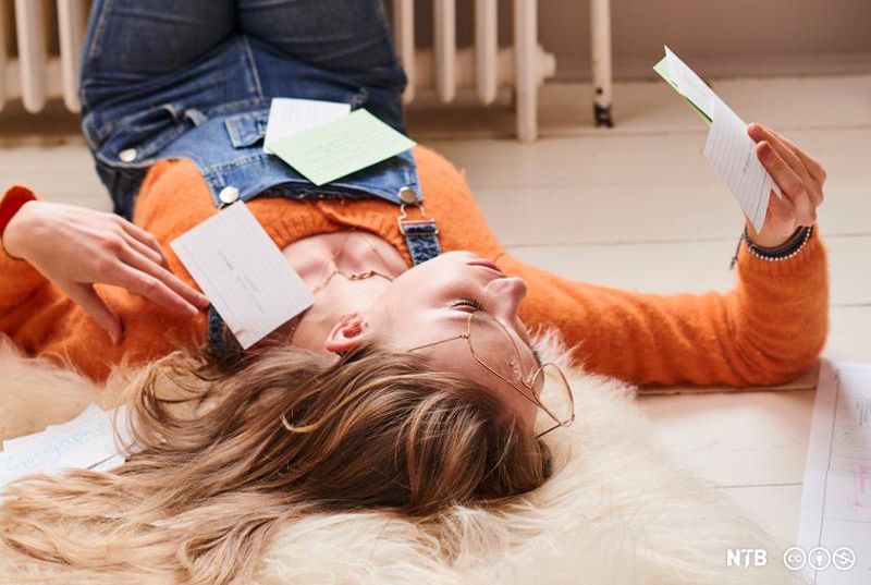 Ei jente kledd i snikkarbukse og oransje genser ligg på golvet og les lappar med notat. Foto.