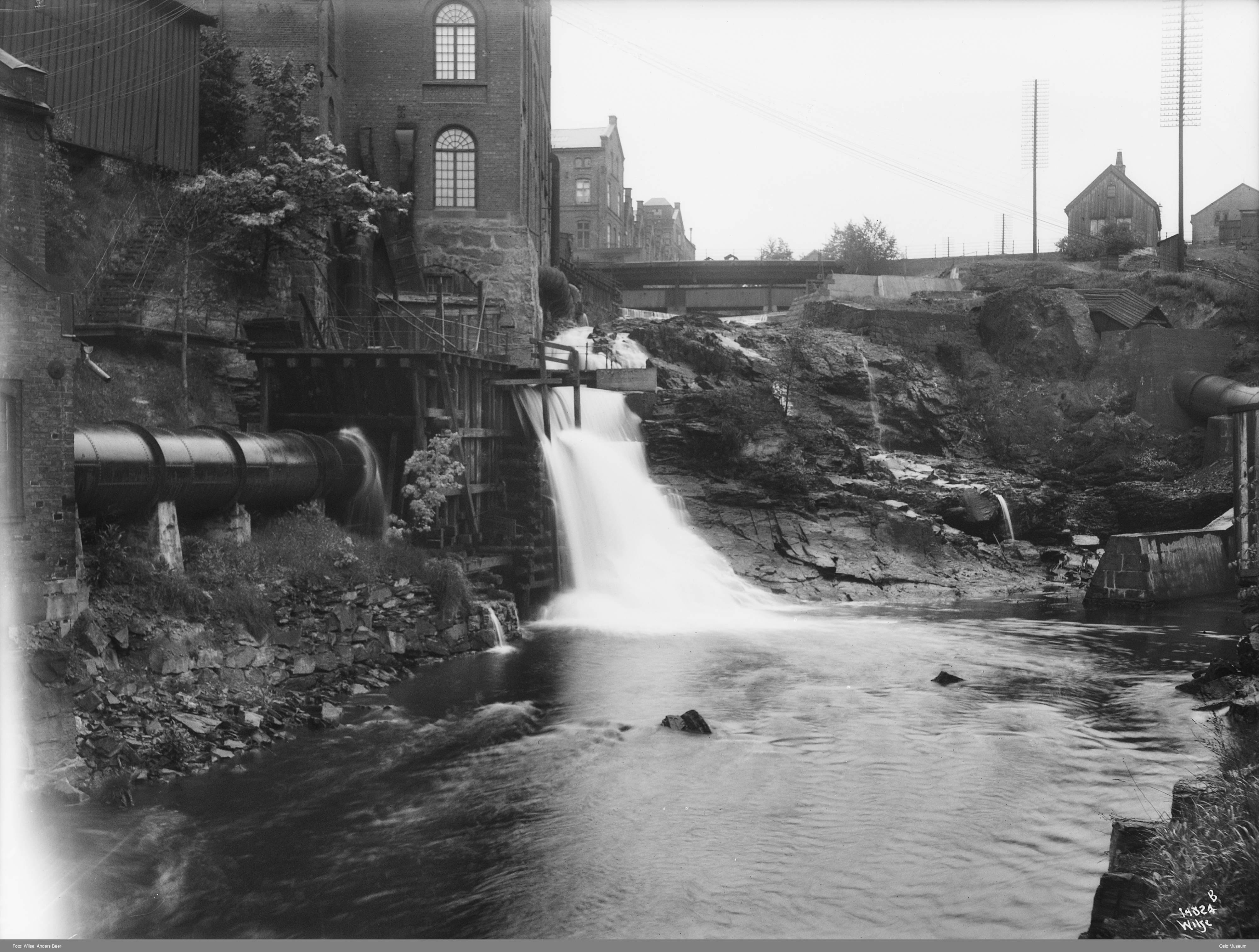 Industribygningar ved fossefall i Akerselva. Ei bru kryssar elva lenger opp. Foto.