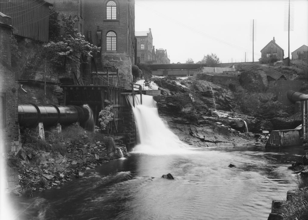 Industribygningar ved fossefall i Akerselva. Ei bru kryssar elva lenger opp. Foto.