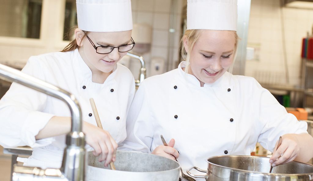 To kokker i rene uniformer jobber ved store gryter. Foto.