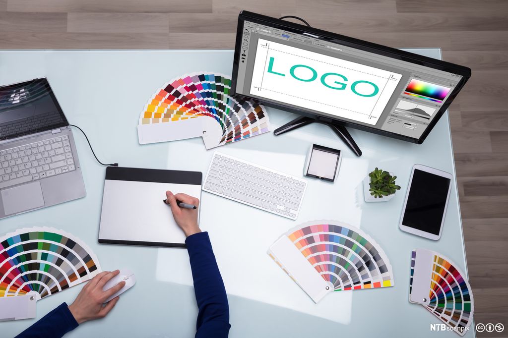 Bilde i fugleperspektiv av arbeidsbordet til en designer. På bordet ligger vifteformede fargepaletter og datautstyr. Vi ser designeren jobbe med tegnebrett og programvare for design. På en dataskjerm står teksten "LOGO". Foto.