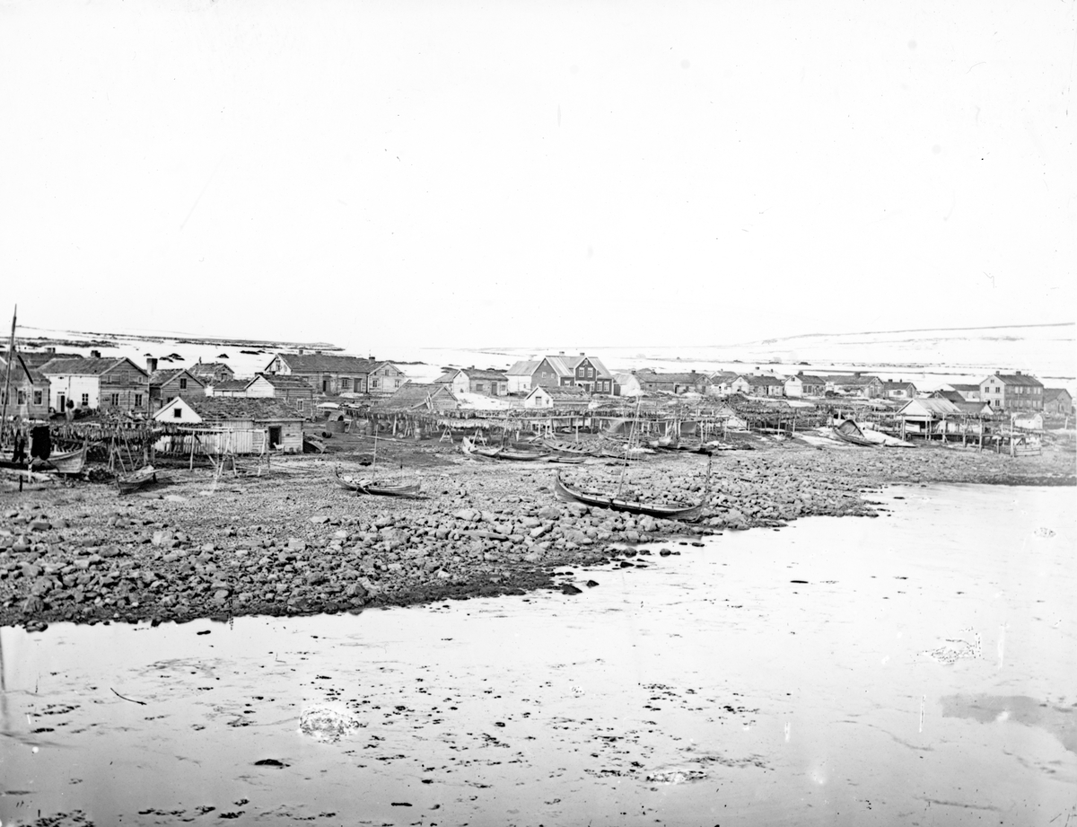 Vadsø på slutten av 1800-tallet. Trehusbebyggelse ved sjøen. Noen få båter i forgrunnen. Foto.