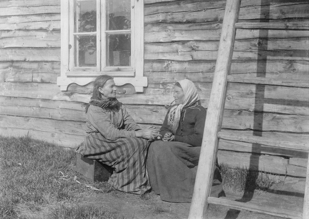 To kvinner i langt skjørt, ei med sjal rundt halsen og ei med skaut, sitter utenfor et trehus. Begge kvinnene røyker pipe. Svart-hvitt foto. 