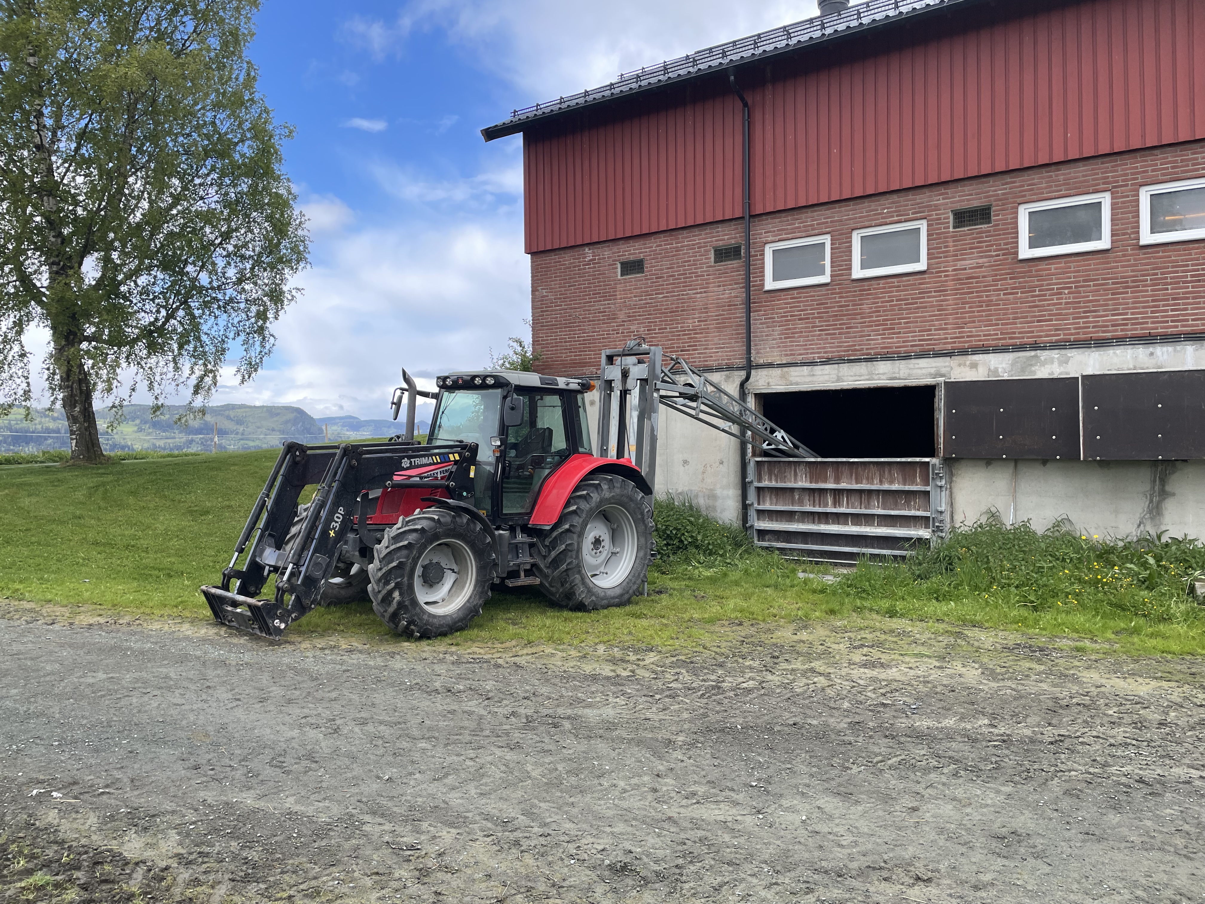 Traktor påmontert redskap bak. Redskapen er ei lang stang som går inn og ned i kjelleren på en fjøsbygning. Foto.