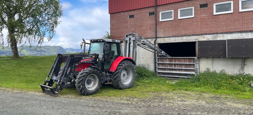 Traktor påmontert redskap bak. Redskapen er ei lang stang som går inn og ned i kjelleren på en fjøsbygning. Foto.