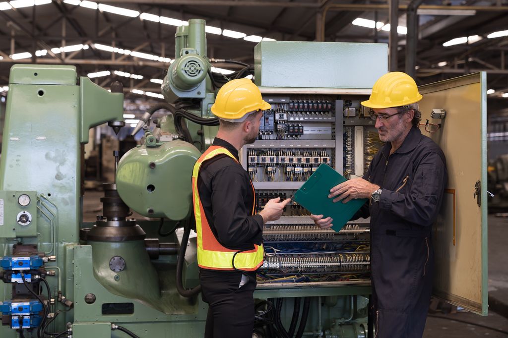 To menn med hjelm inspiserer det elektriske systemet til en CNC-maskin i en fabrikk. Foto.